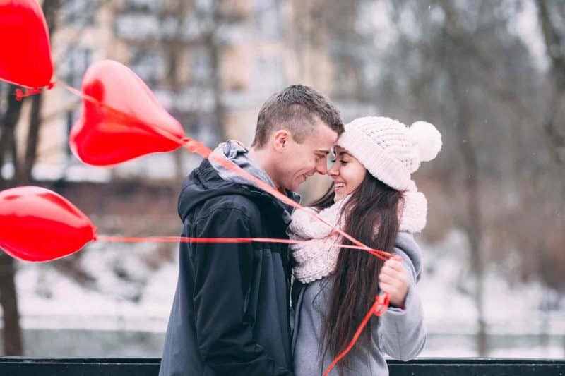 A couple in love standing close with nose touching and red heart balloons in hand